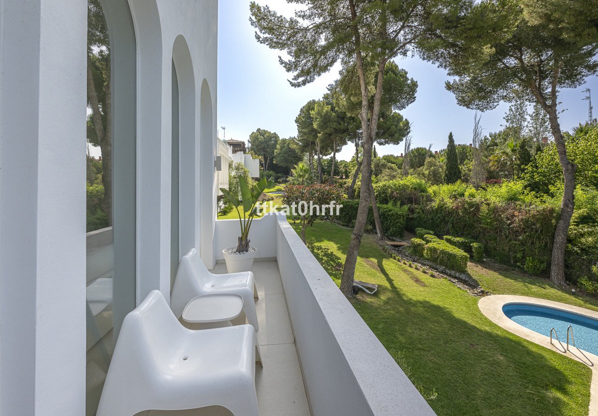 A balcony with two white chairs overlooking a lush green garden with trees and a blue swimming pool.