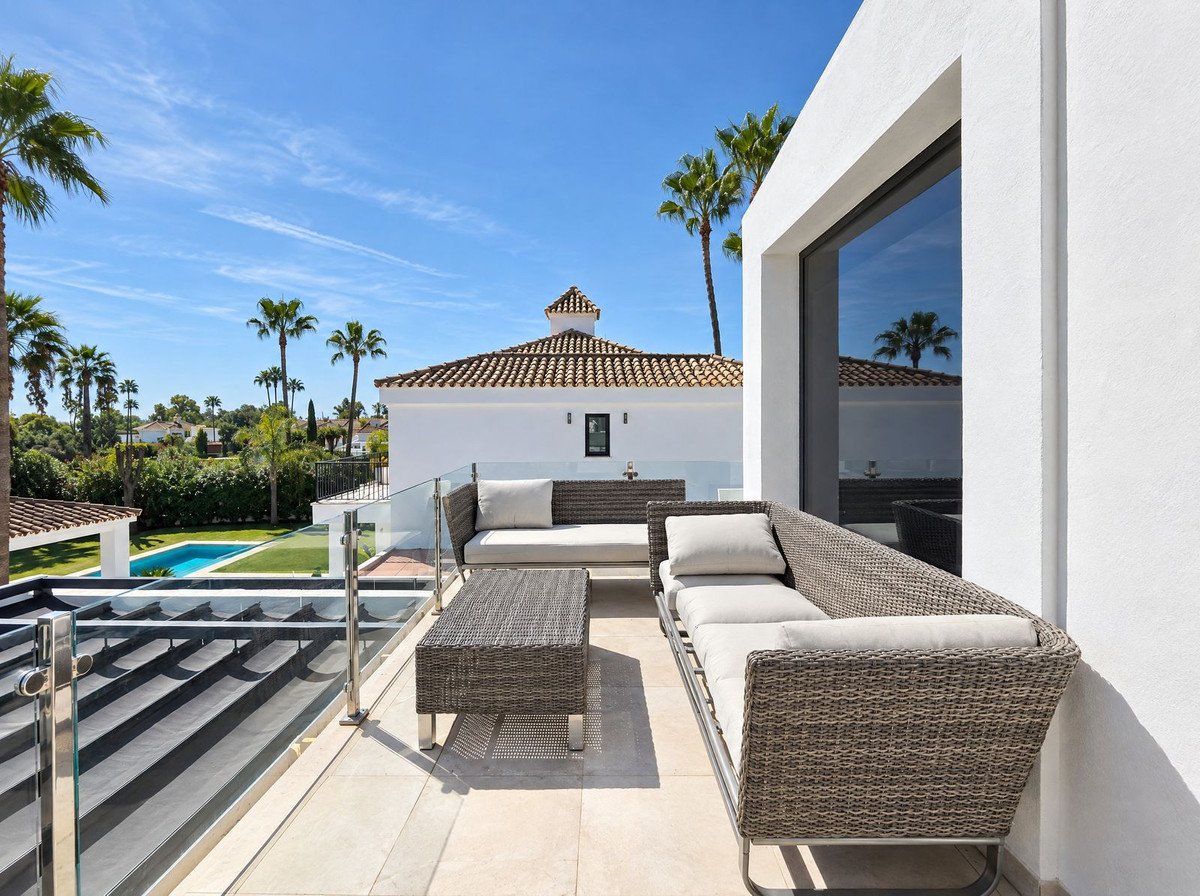 Modern terrace with wicker seating, coffee table, glass railings, overlooking a swimming pool and palm trees.