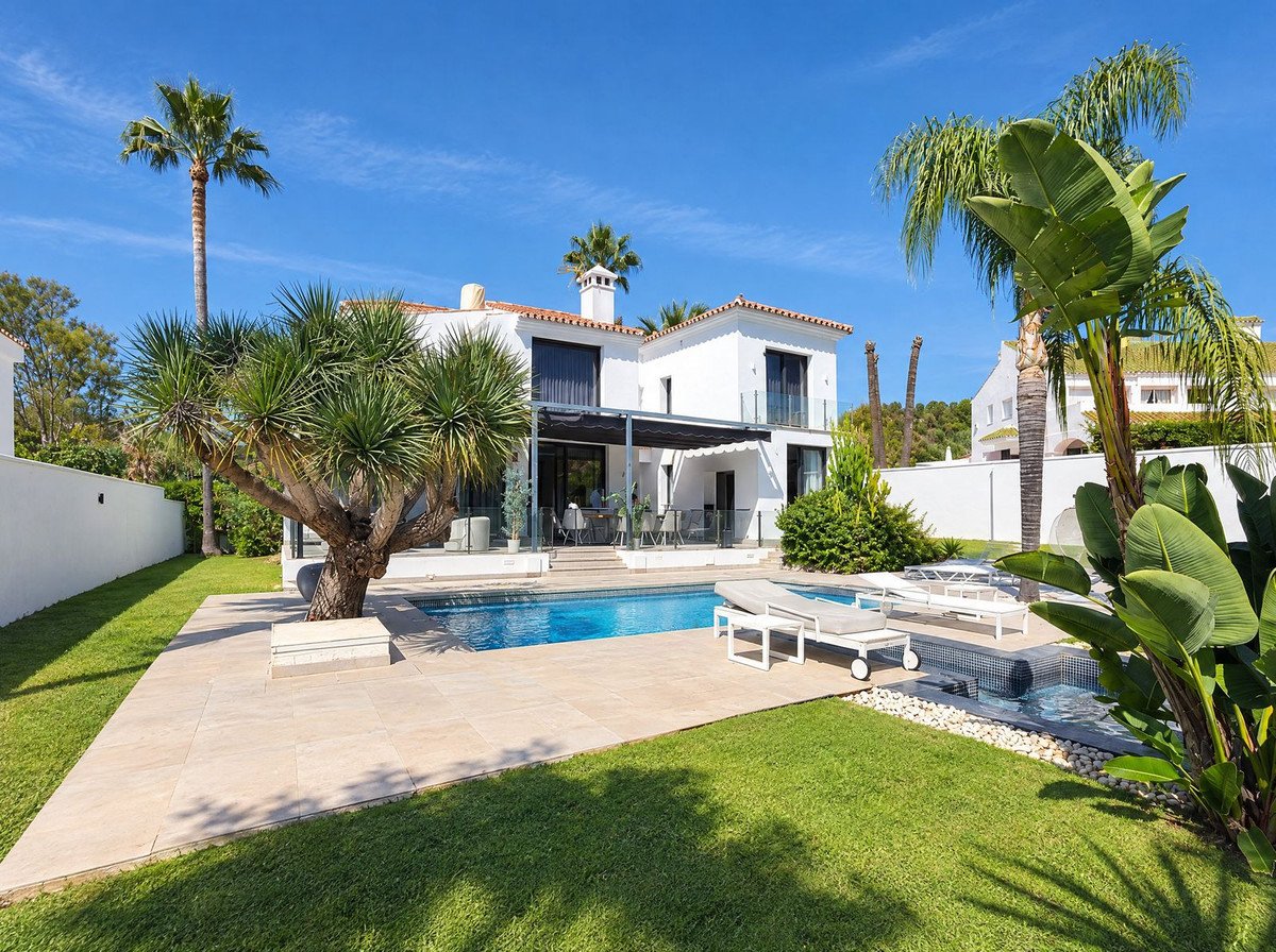 A modern white villa with a blue swimming pool, sun loungers, and lush green garden under a clear blue sky.