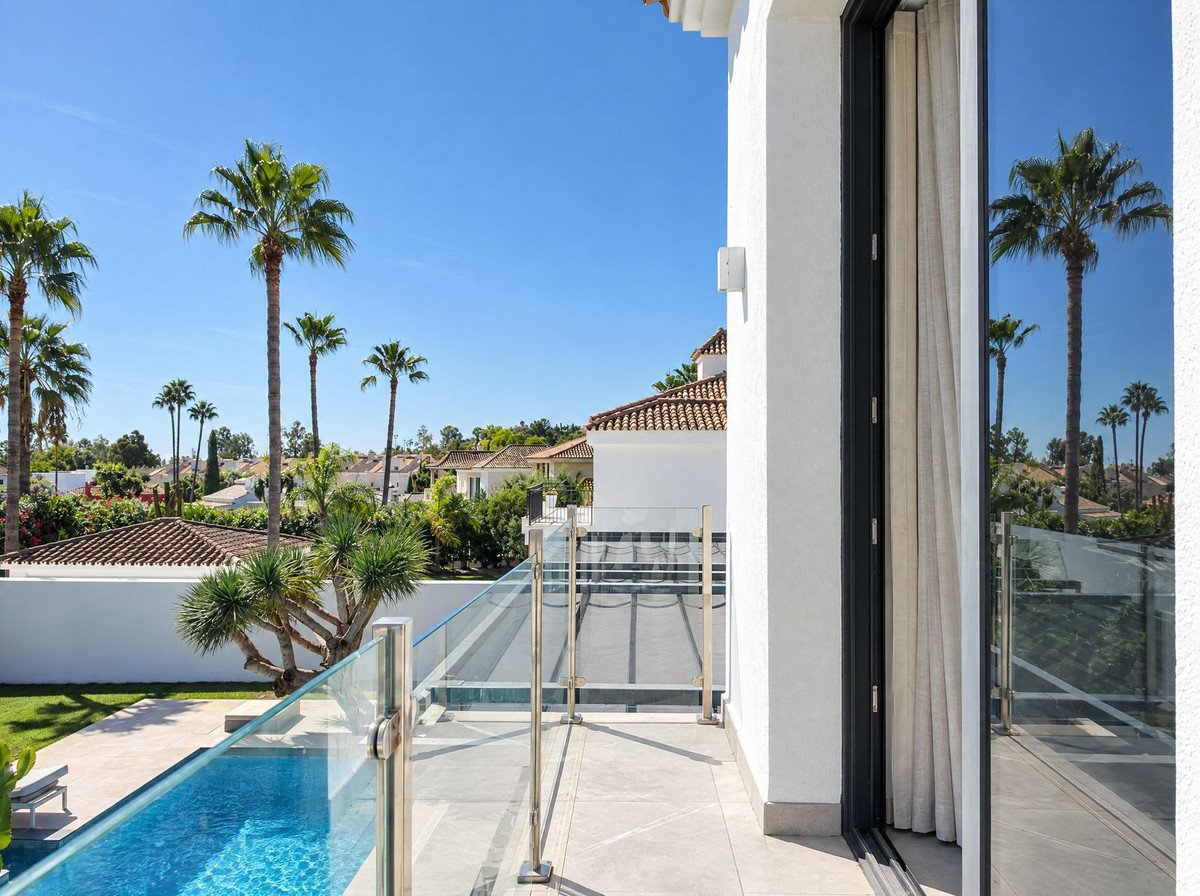 Modern balcony with glass railing overlooking a pool and palm trees with a blue sky.