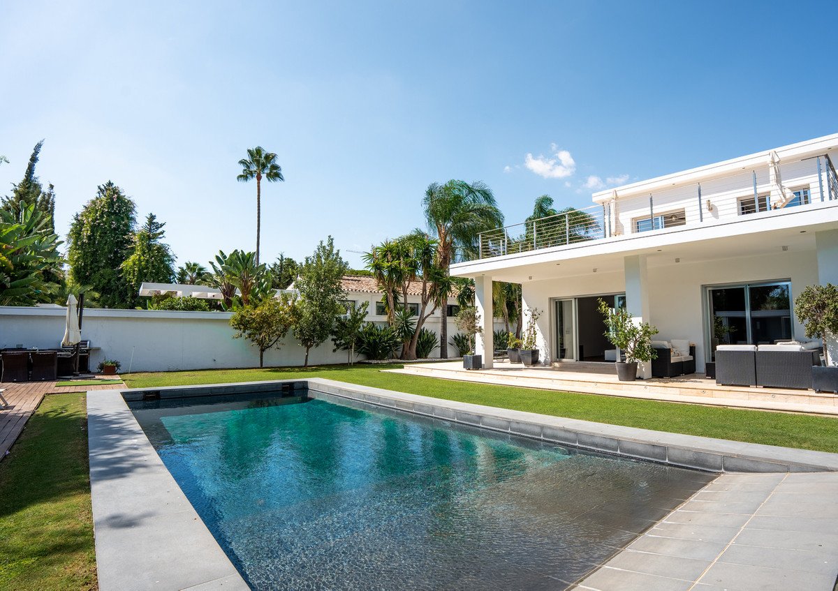 Modern white villa with a swimming pool, lawn, terrace, and tropical garden under a clear blue sky.