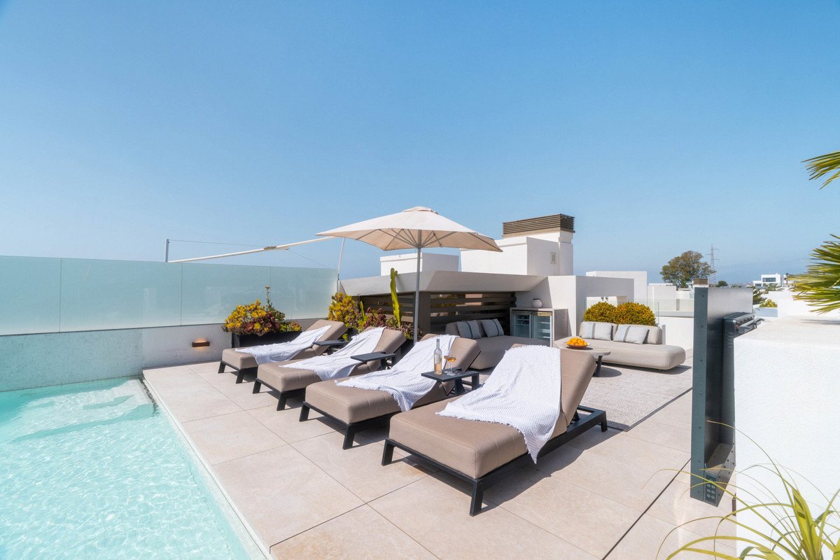 A luxurious rooftop terrace with a pool, lounge chairs, a sofa, and an outdoor bar under a clear blue sky.