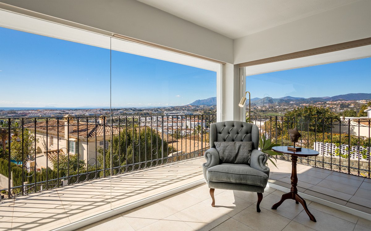 Armchair with a side table on a balcony with a panoramic view of a town and mountains under a clear blue sky.