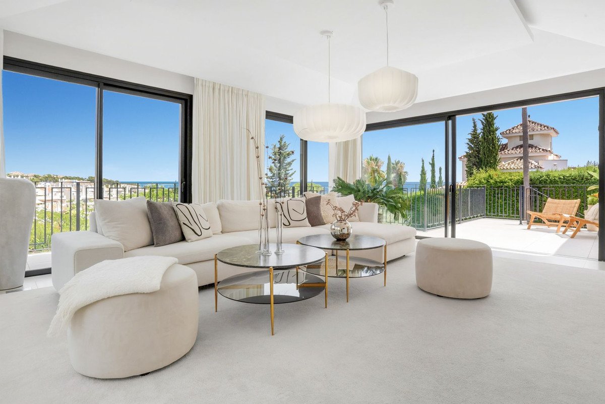 Modern living room with large windows, comfortable sofa, coffee tables, and a balcony with sea and garden views.