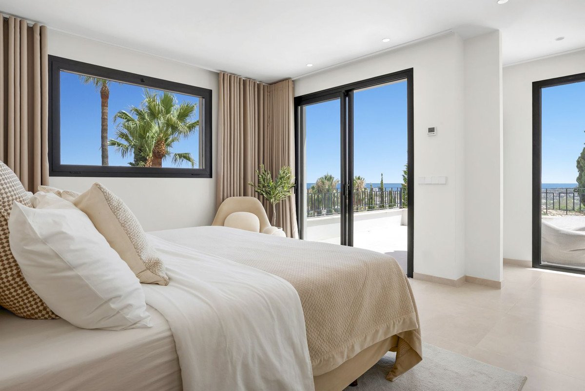 Bedroom with a large bed, large windows and sliding doors offering a view of the sea, balcony, and palm trees.