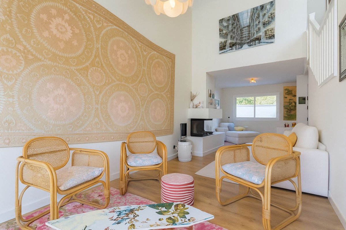 A living room with rattan chairs, a fireplace, and a large tapestry on the wall. Large windows offer natural light.