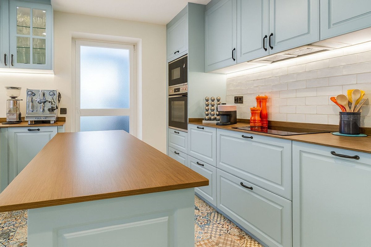 A modern kitchen with light blue cabinets, a wooden island, integrated appliances, and a white subway tile backsplash. Natural light streams in.