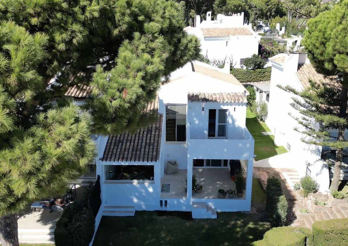 A modern white villa with large windows, a covered patio, and a green garden surrounded by trees.