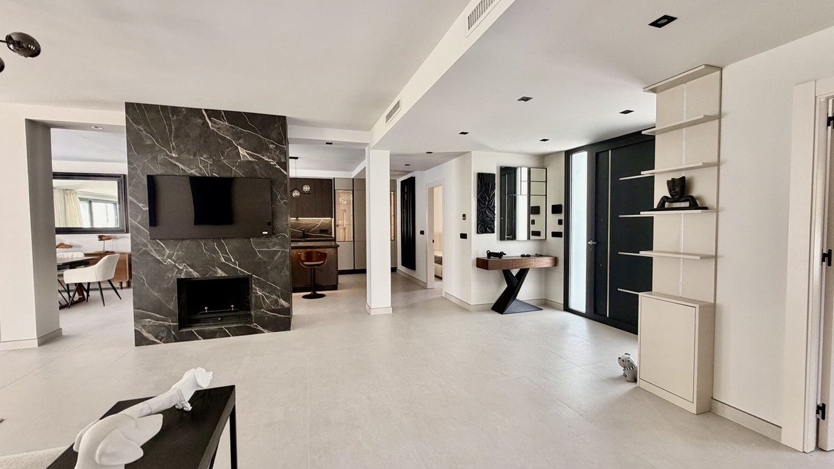 A modern open-plan living room with a marble fireplace, mounted TV, and view into a contemporary kitchen.
