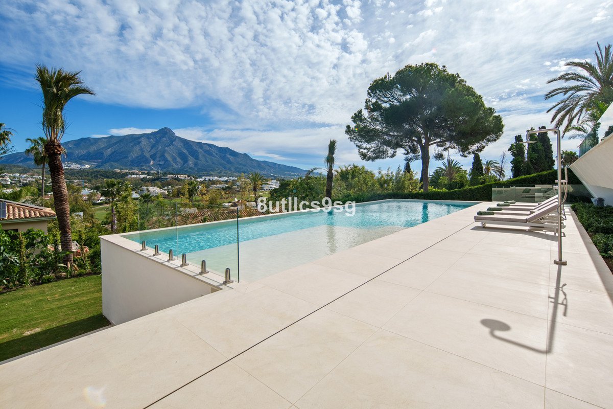 Infinity swimming pool on a terrace with lounge chairs, overlooking a mountain range and lush greenery under a blue sky.