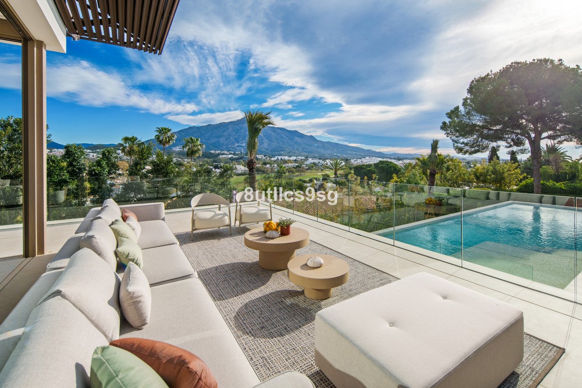 A modern terrace with a large sofa, coffee tables, and swimming pool, offering a panoramic view of mountains and trees.