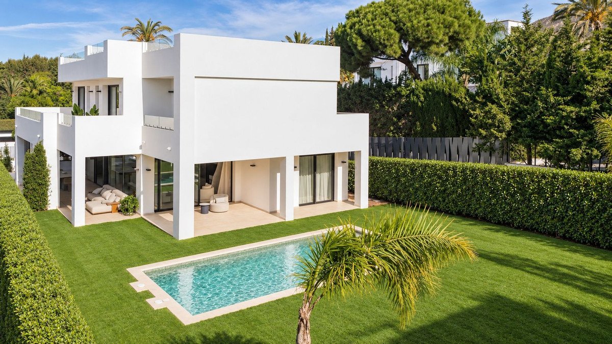 A modern white villa with a swimming pool, manicured lawns, and lush green hedges on a sunny day.