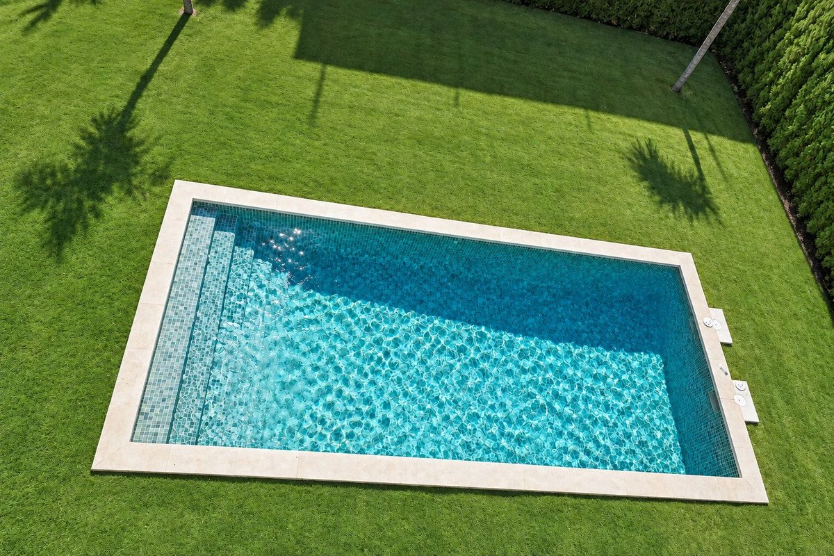 Rectangular swimming pool with tiled steps, surrounded by green grass and palm tree shadows.