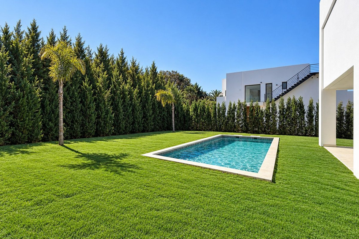 A rectangular swimming pool set in a lush green lawn, surrounded by tall cypress trees and a modern white house with outdoor stairs.