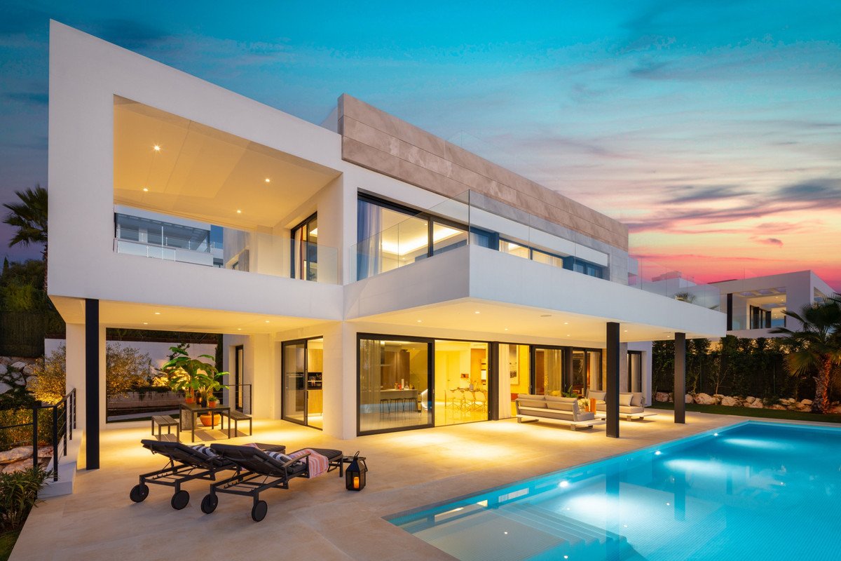 Modern villa with a swimming pool and lounge area at sunset, featuring large windows and balconies.