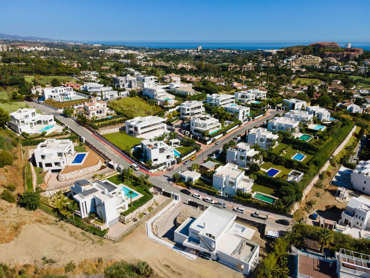Aerial view of modern white villas with swimming pools nestled in a green hillside, overlooking the blue sea under a clear sky.