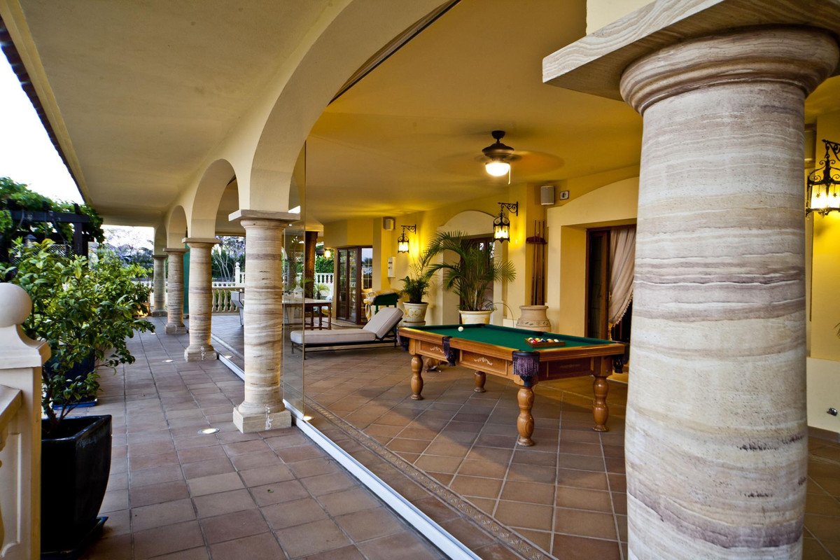 Covered patio featuring a pool table and lounge area with arched columns and lush greenery visible greenery.