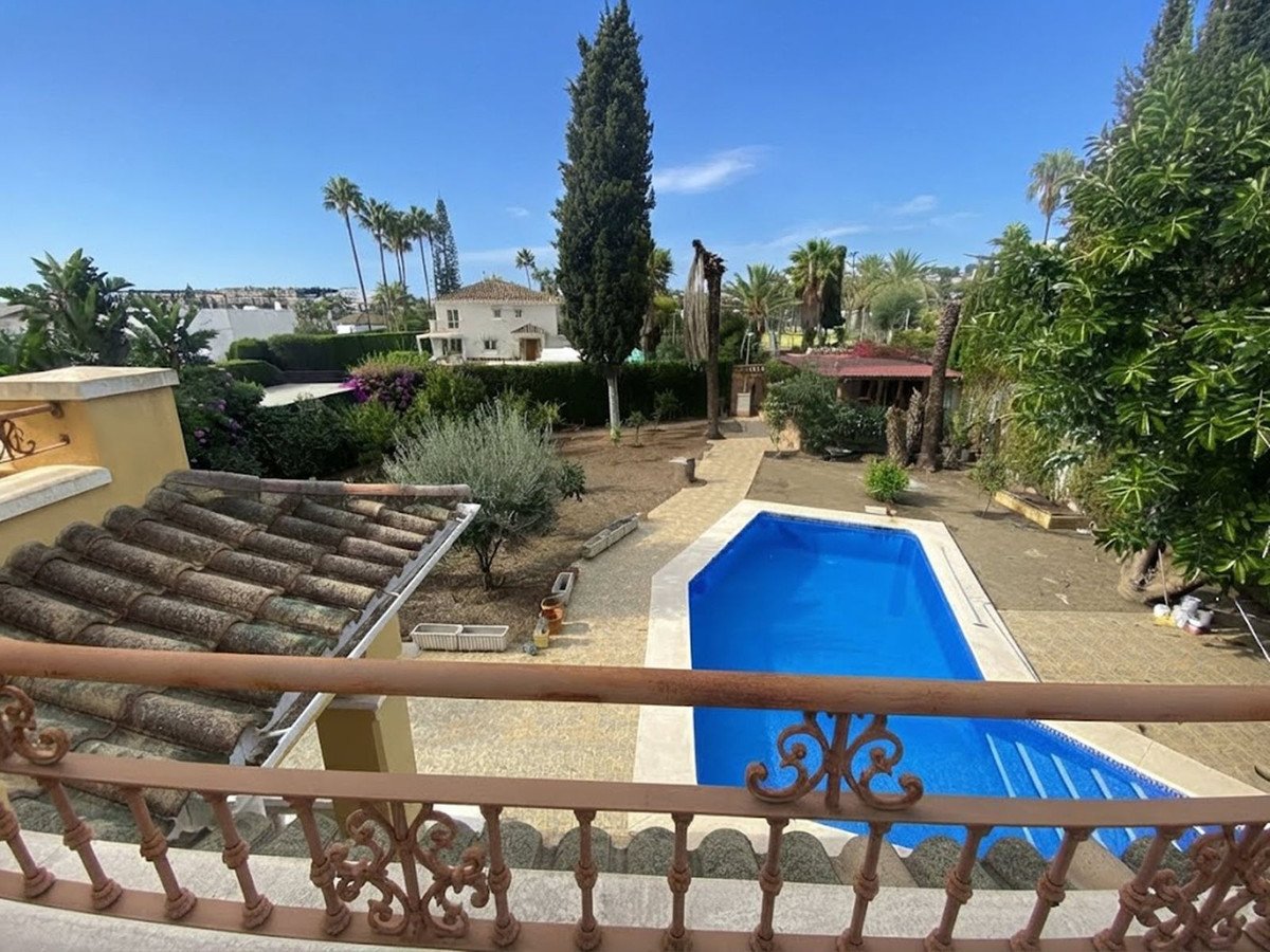 A bright blue swimming pool in a garden with palm trees and a villa in the background under a clear blue sky.