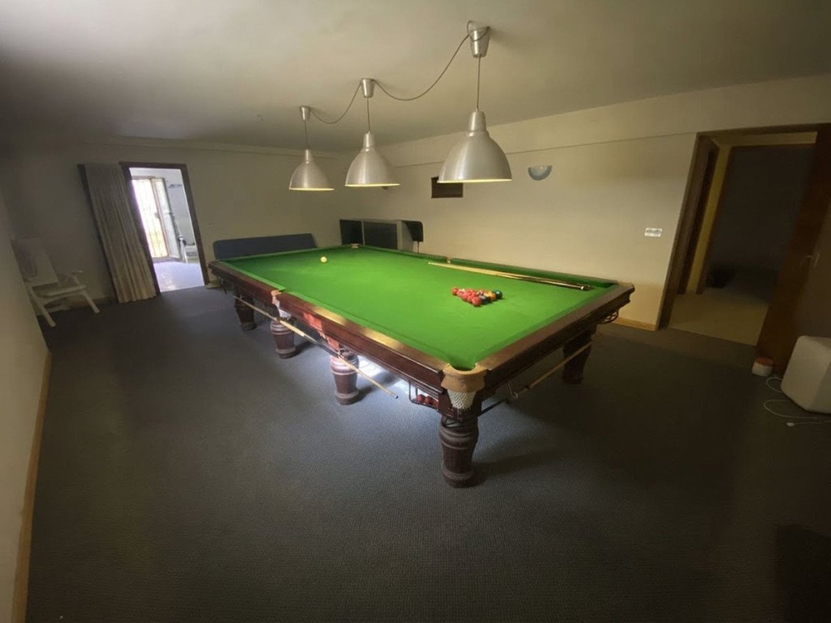 A spacious room featuring a snooker table with balls and a cue, illuminated by pendant lights. A doorway leads outside.
