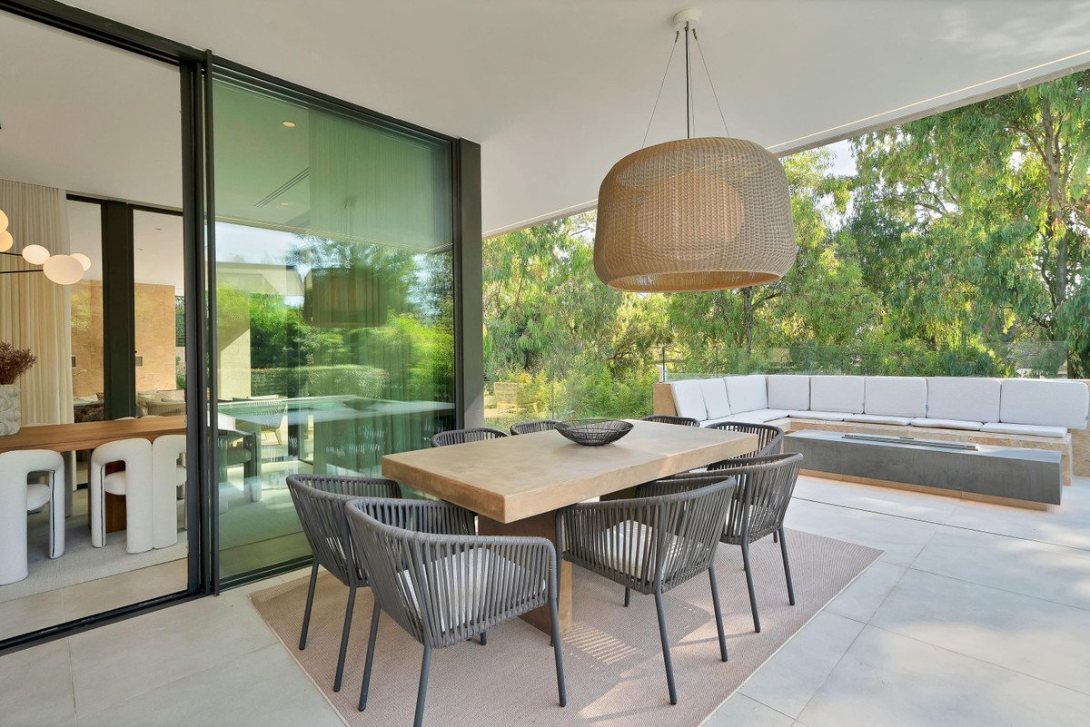 Modern outdoor dining area with wooden table, wicker chairs, and a view of greenery through large glass doors.