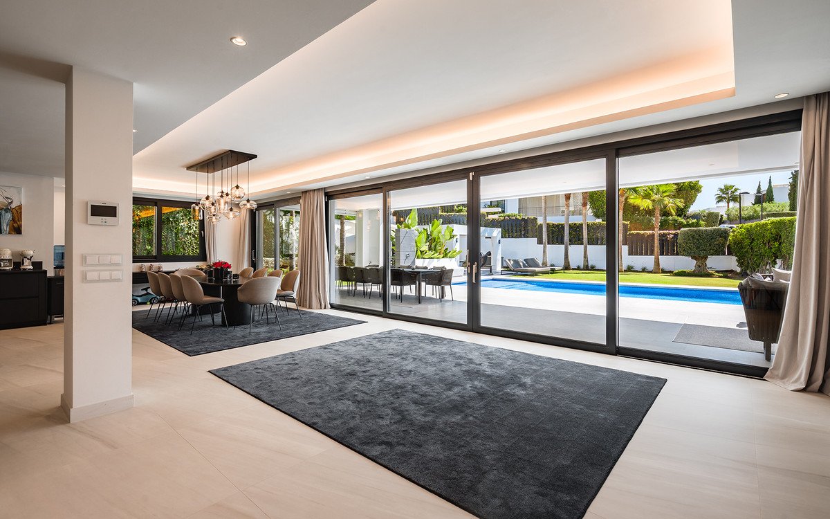 Modern dining area with a large table and chairs, opening to a patio with a swimming pool and garden.