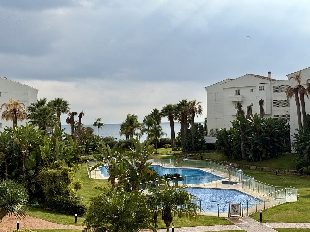 A resort complex with a swimming pool surrounded by lush tropical gardens and palm trees, overlooking the sea on a cloudy day.