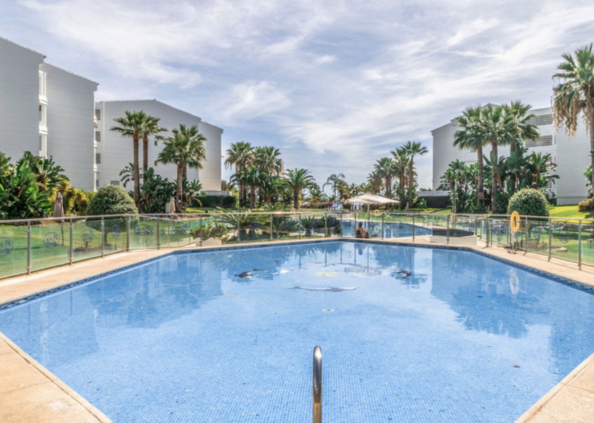 A clear blue swimming pool surrounded by palm trees and resort buildings under a cloudy sky.