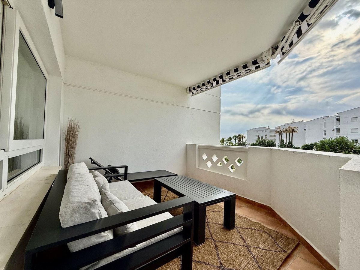 A furnished terrace with a sofa, table, and a view of buildings and palm trees under a cloudy sky.