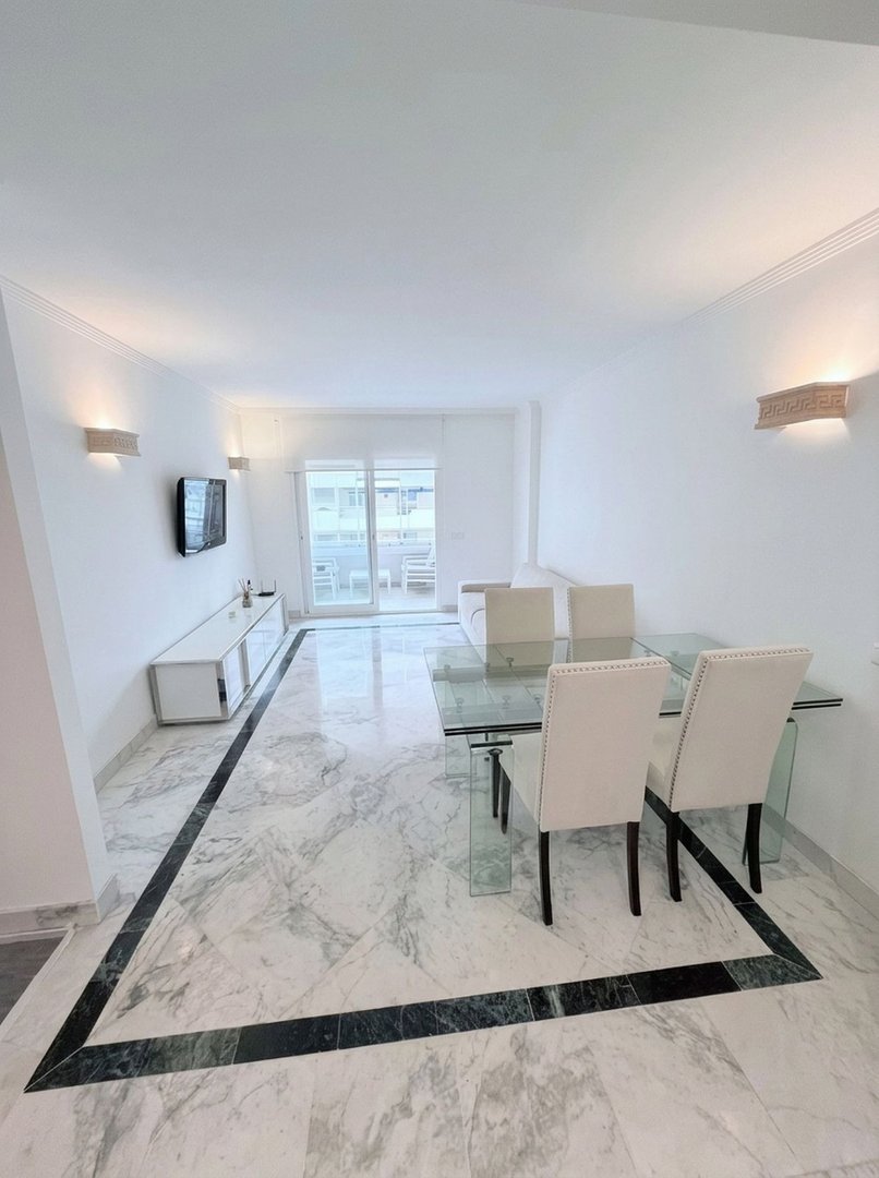 Spacious living and dining area with a glass table, modern furniture, and a balcony visible through large sliding doors.