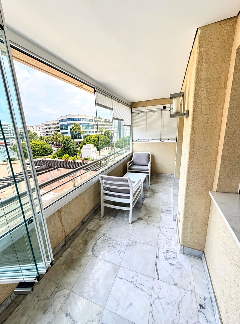 Enclosed balcony with modern furniture and marble floors, offering a view of buildings and trees.