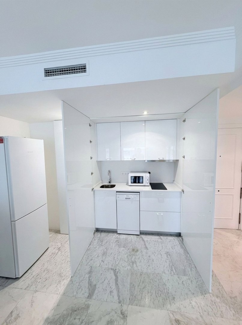 A compact, modern white kitchen featuring integrated appliances, a microwave, and sleek cabinetry, with marble-patterned flooring.
