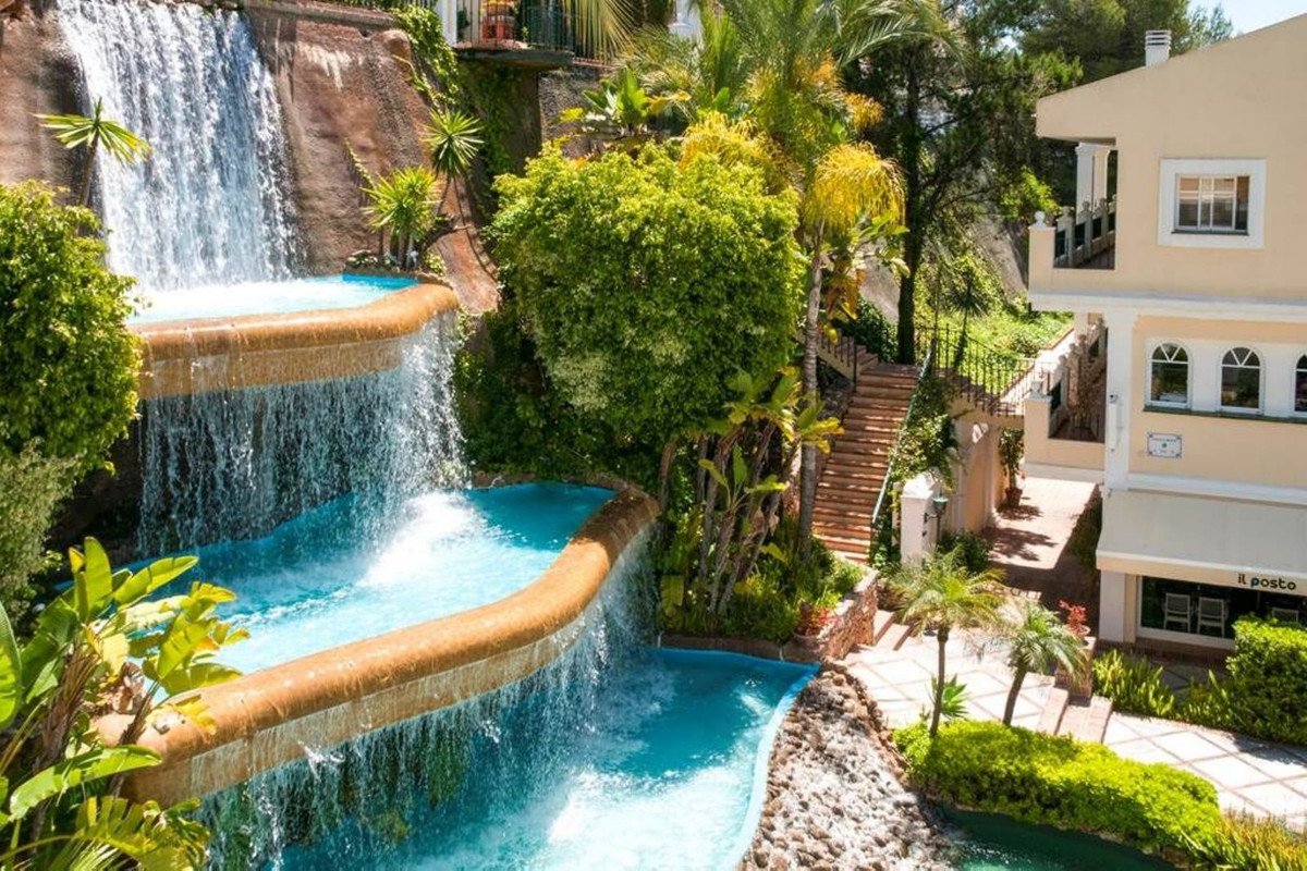 Luxurious multi-tiered waterfall cascading into pools, surrounded by lush tropical greenery and a building.