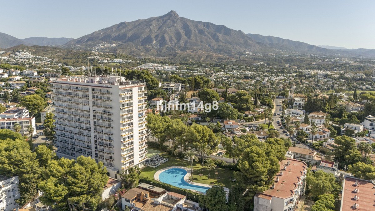 Aerial view of a modern apartment building with a pool and lush greenery, overlooking a town and mountains.