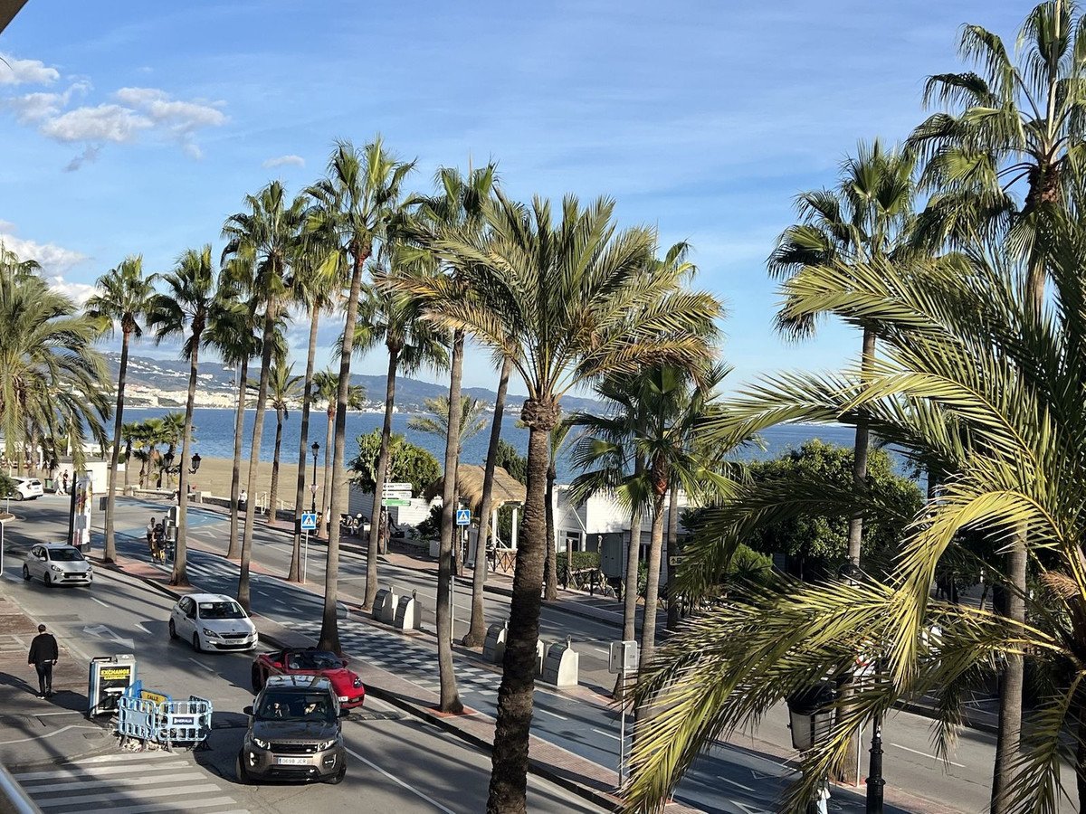 A street lined with palm trees leading to a view of the blue sea and distant hills under a clear sky.