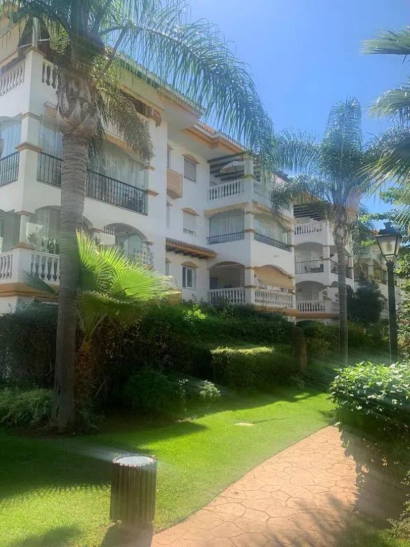 A white apartment building with balconies surrounded by lush green gardens and palm trees on a sunny day.