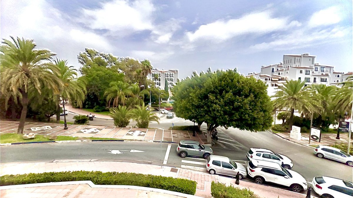 Street view with palm trees, parked cars, and white residential buildings under a partly cloudy sky.