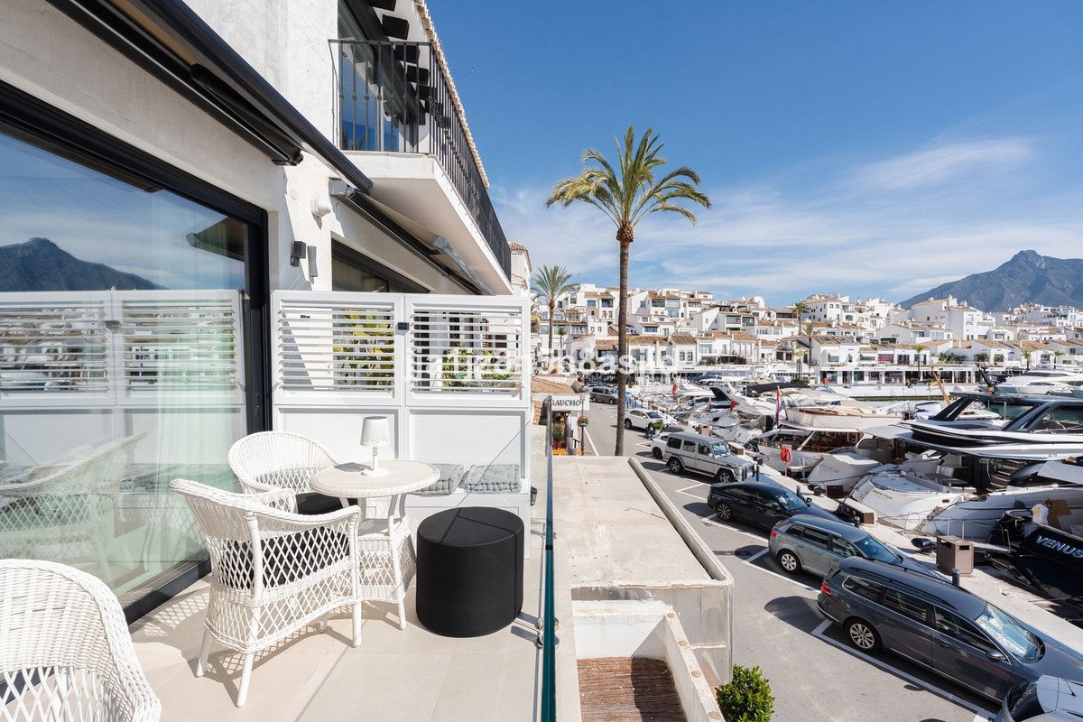 A white wicker seating area on a balcony overlooking a bustling marina with boats and white buildings.