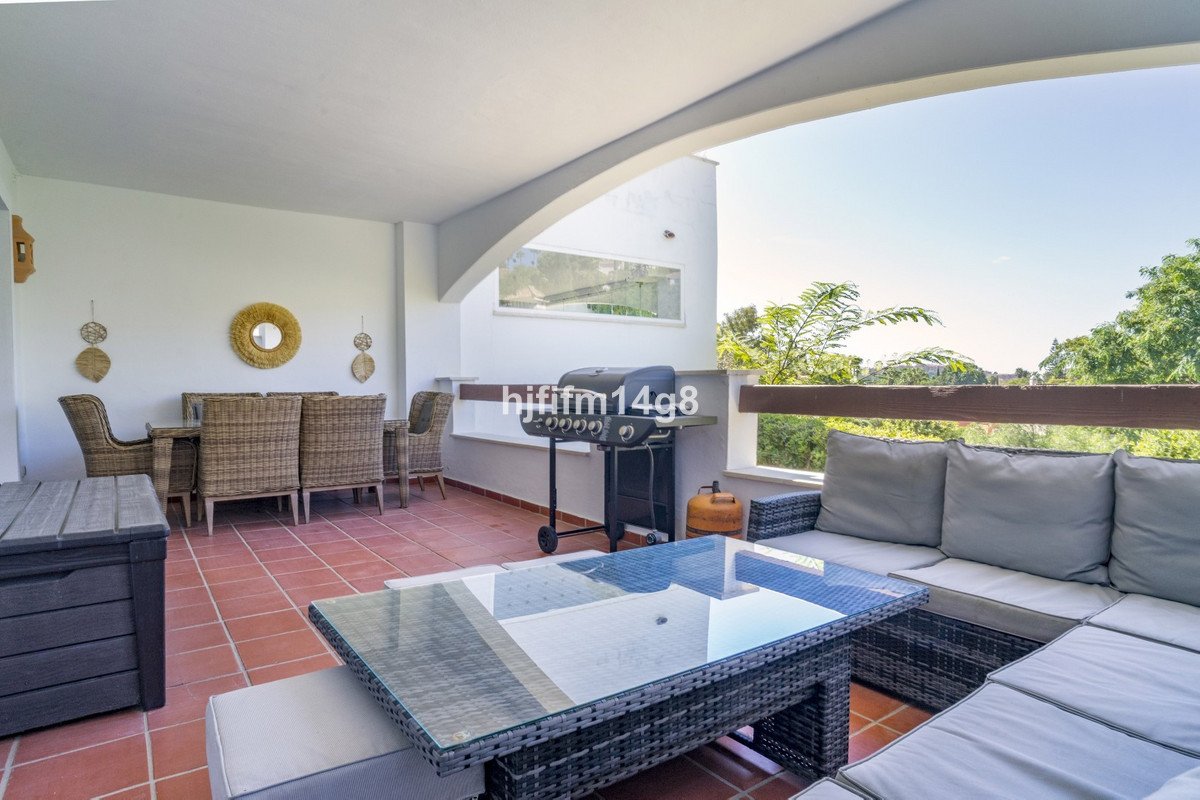 Covered patio with dining set, lounge sofa, coffee table, and grill, overlooking greenery.