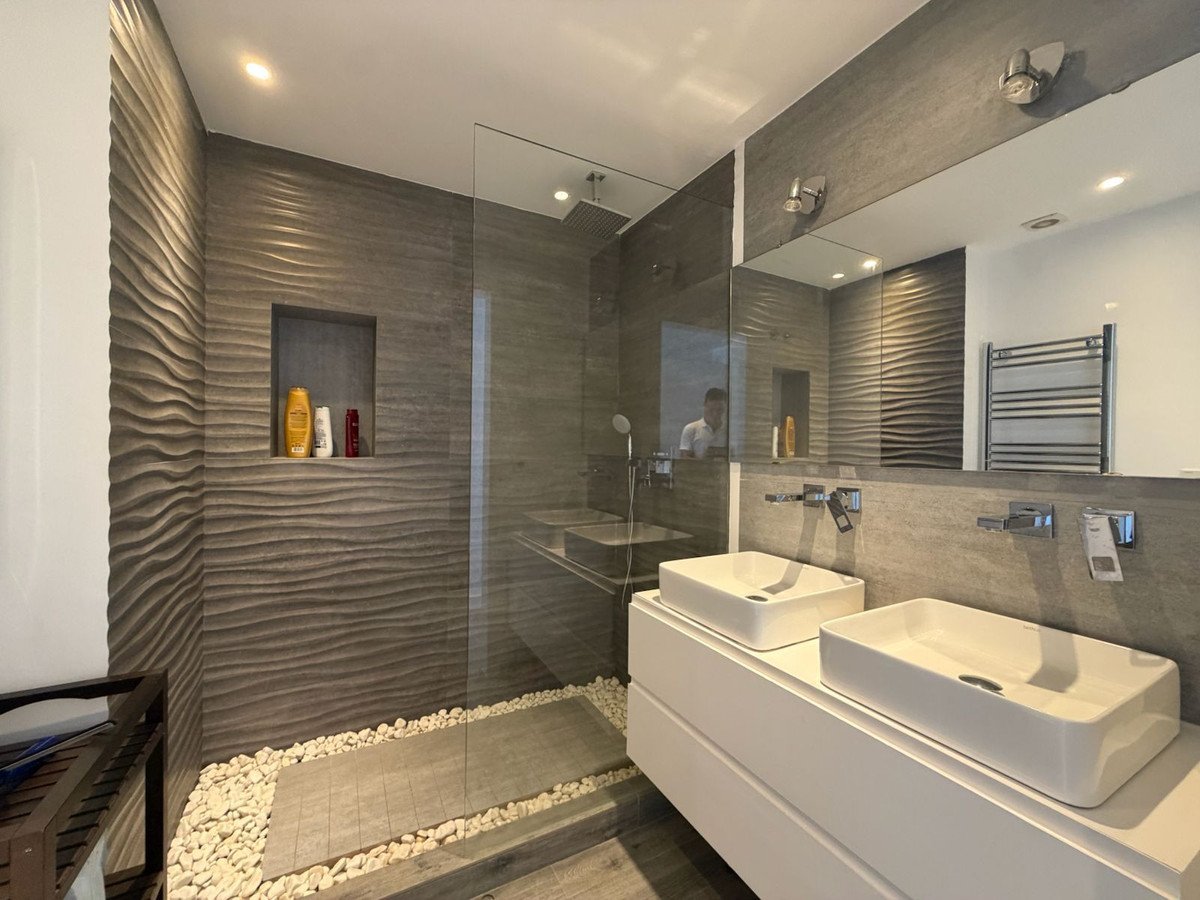 A modern bathroom features a double vanity with vessel sinks, a walk-in shower with wave tile, and a pebble floor detail.