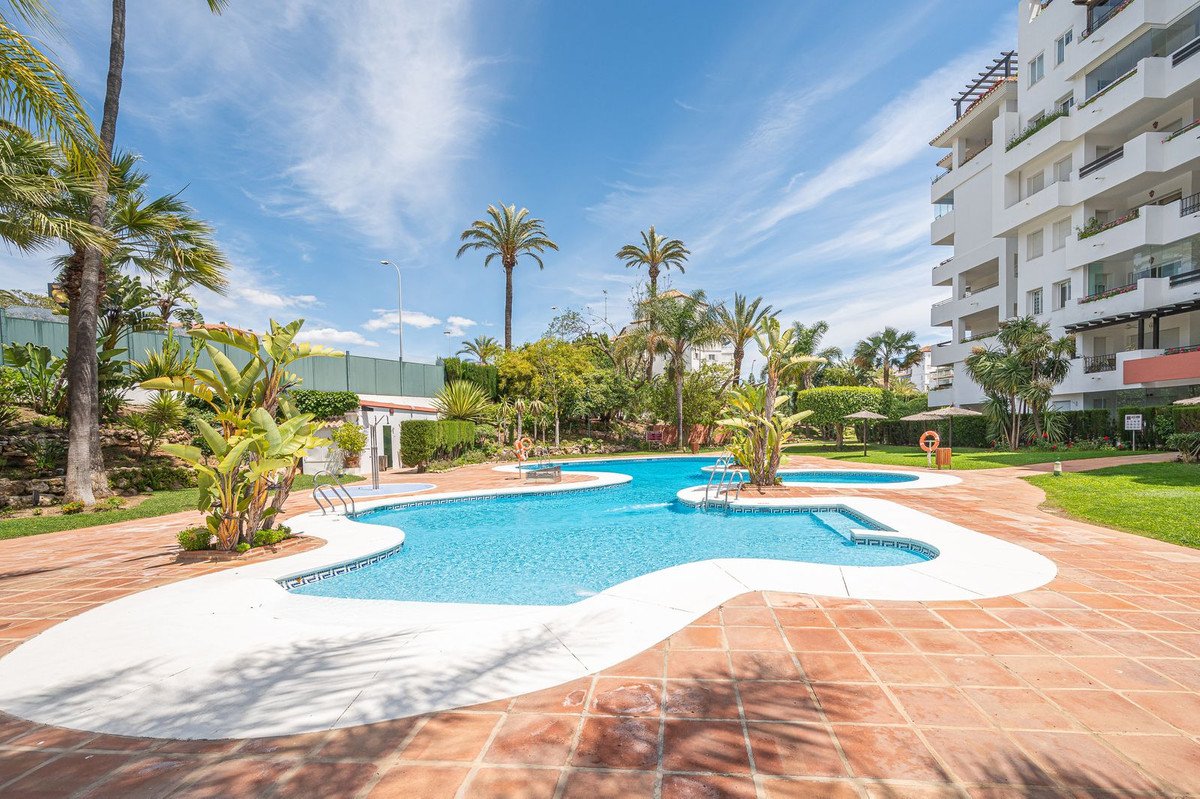 A resort-style swimming pool surrounded by lush greenery and palm trees, with a modern apartment building in the background.