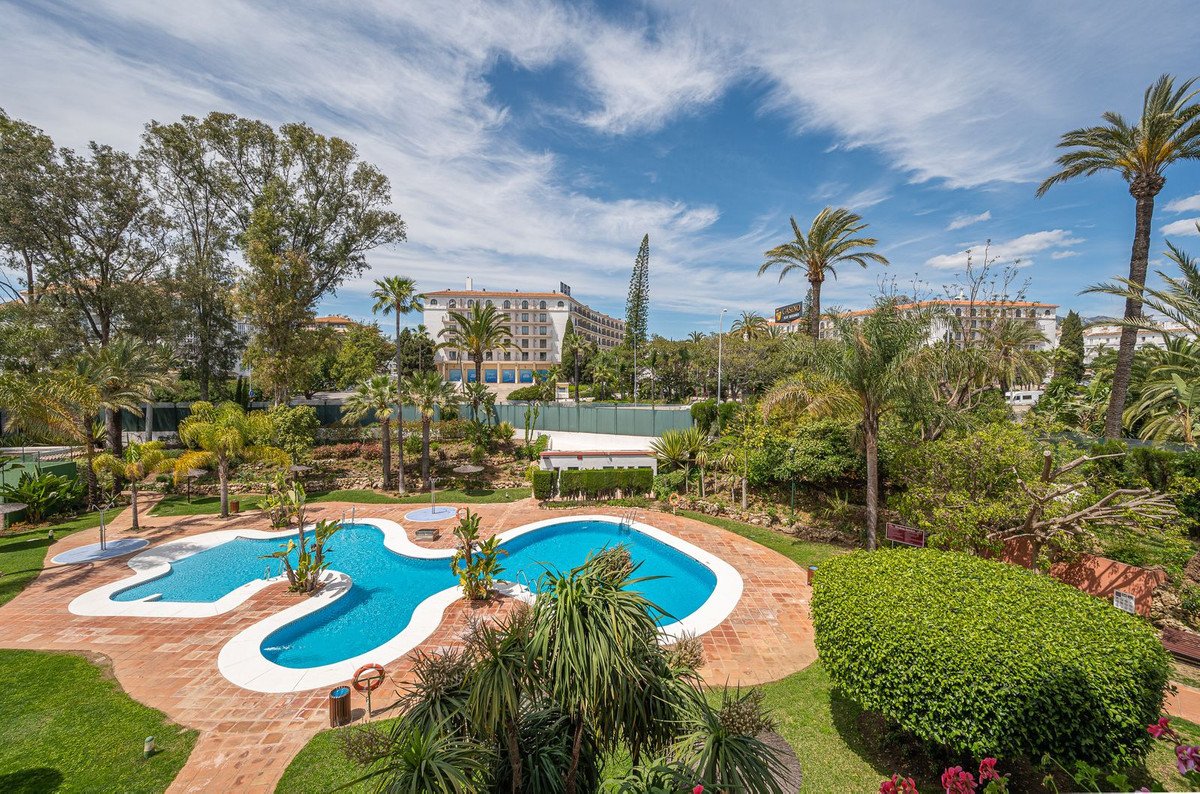 A resort-style swimming pool surrounded by lush green gardens and palm trees, with a hotel building in the background.