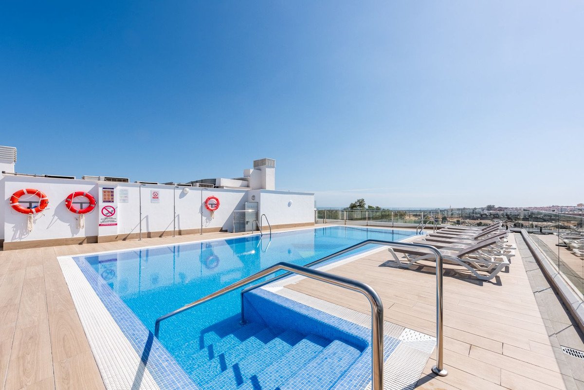 A rooftop swimming pool with blue water, surrounded by wooden decking and loungers, under a clear blue sky.