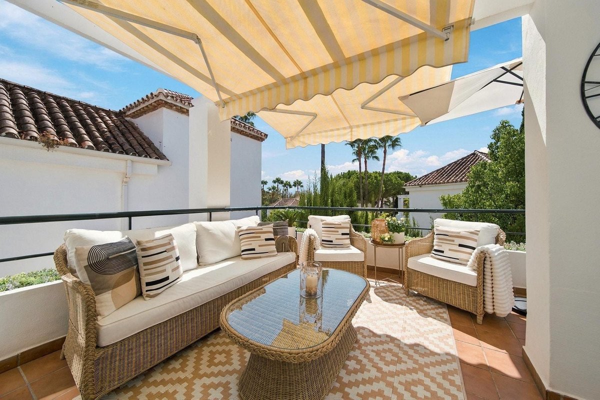 Comfortable outdoor seating on a terrace with a glass coffee table and a view of palm trees under a striped awning.