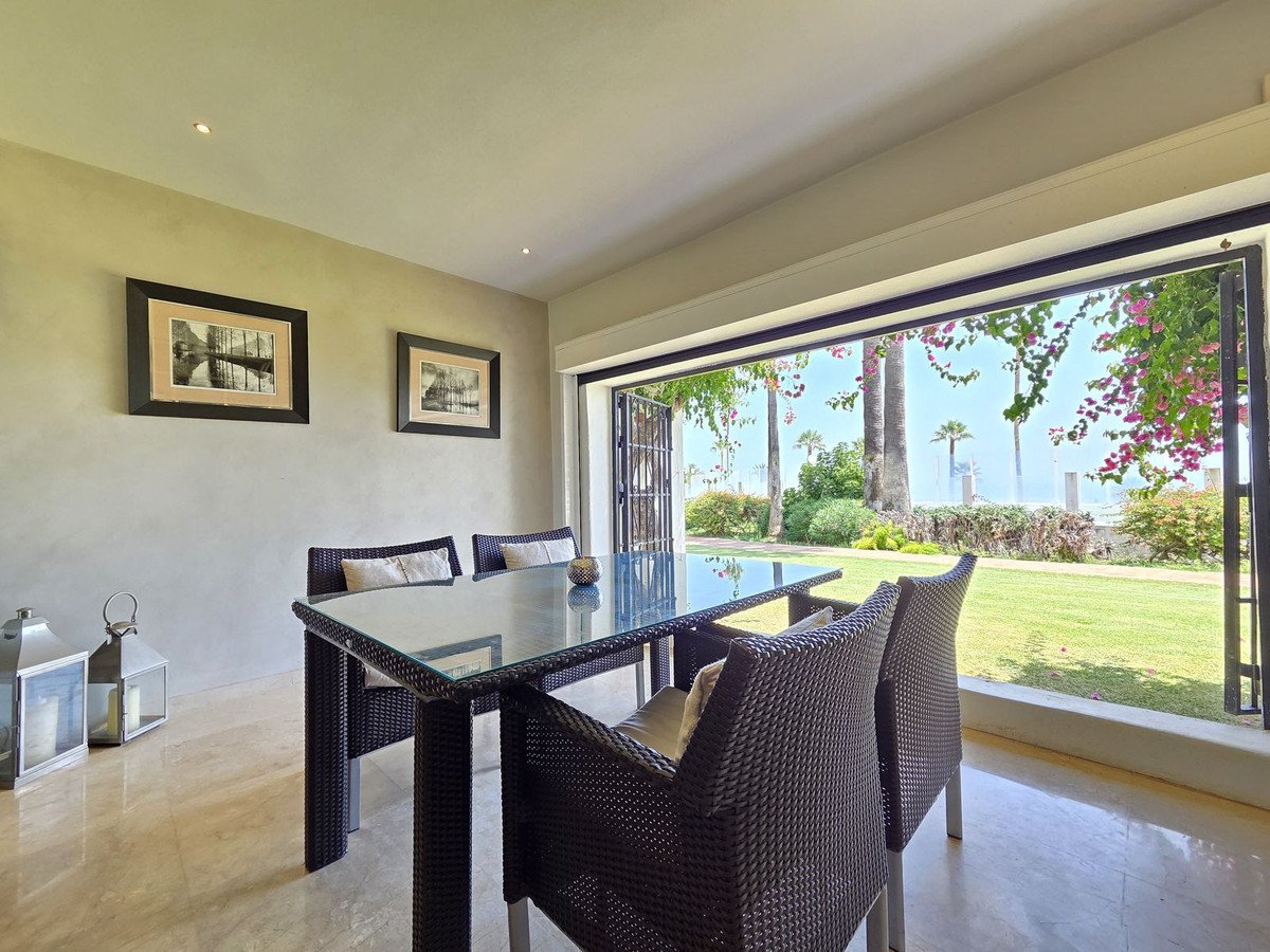 Dining area with glass table and chairs opening to a garden with ocean view and palm trees.