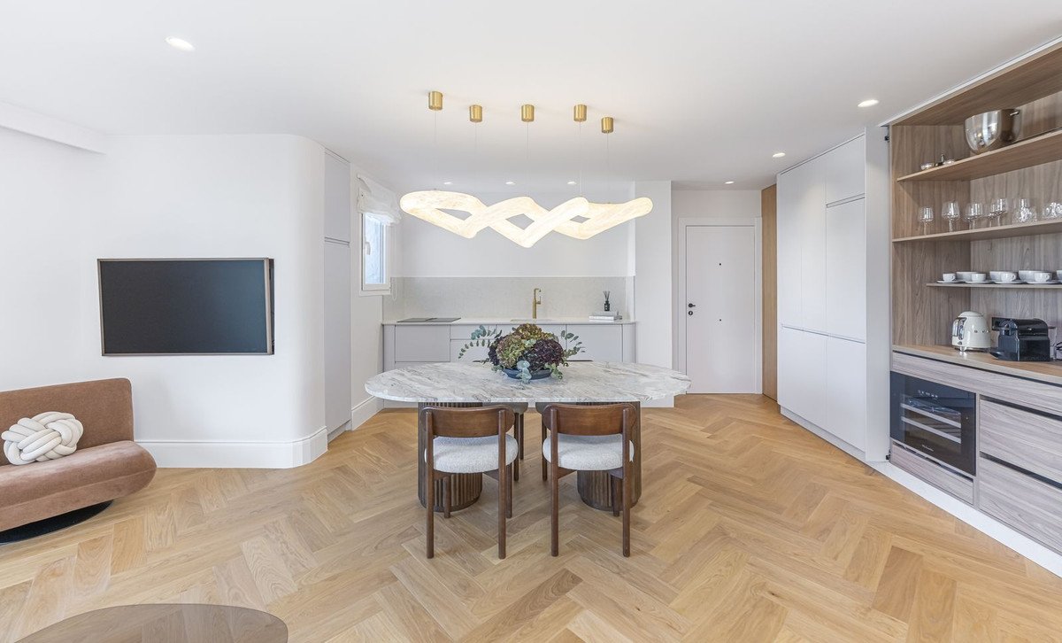 Modern open-plan living, dining, and kitchen area with herringbone wood flooring, marble table, and contemporary lighting.