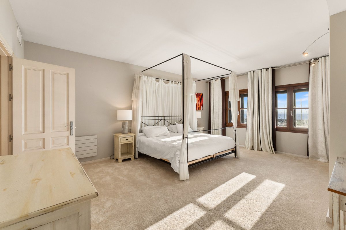 Spacious bedroom with four-poster bed, white linens, and large windows offering a sea view.