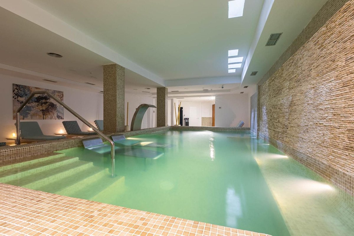 Indoor spa pool with gentle waterfall feature, integrated steps, lounge chairs, and tiled stone wall.