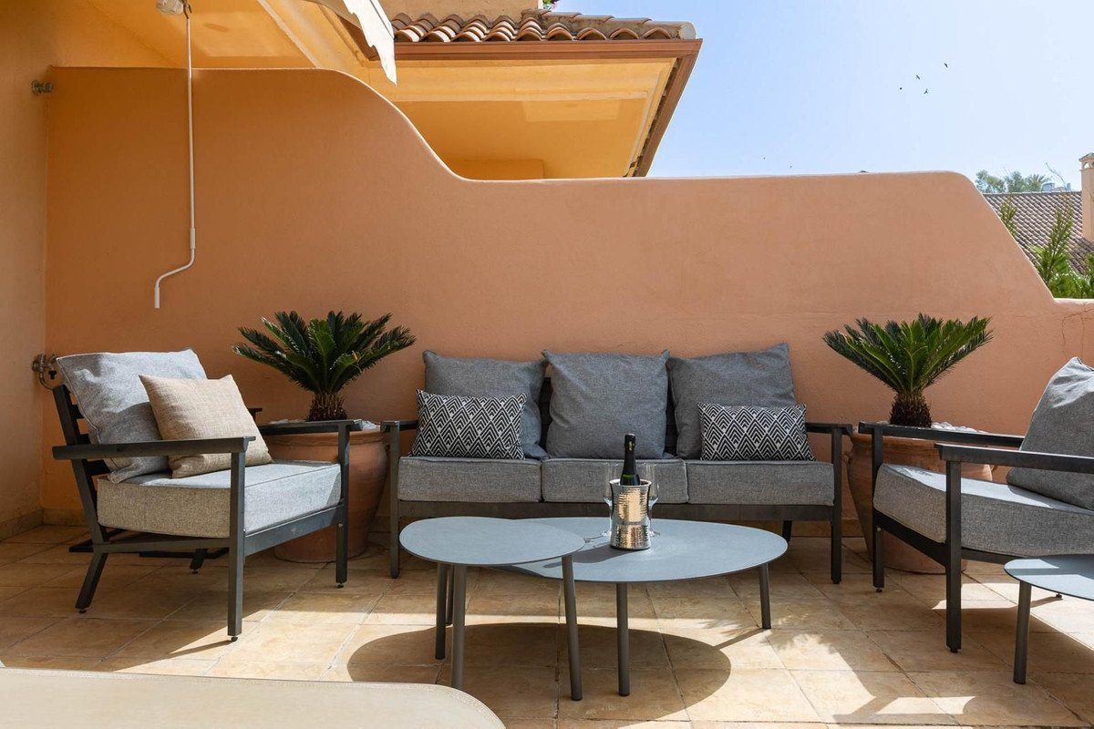 Outdoor terrace with grey sofa, chairs, coffee table, and plants on a sunny day.