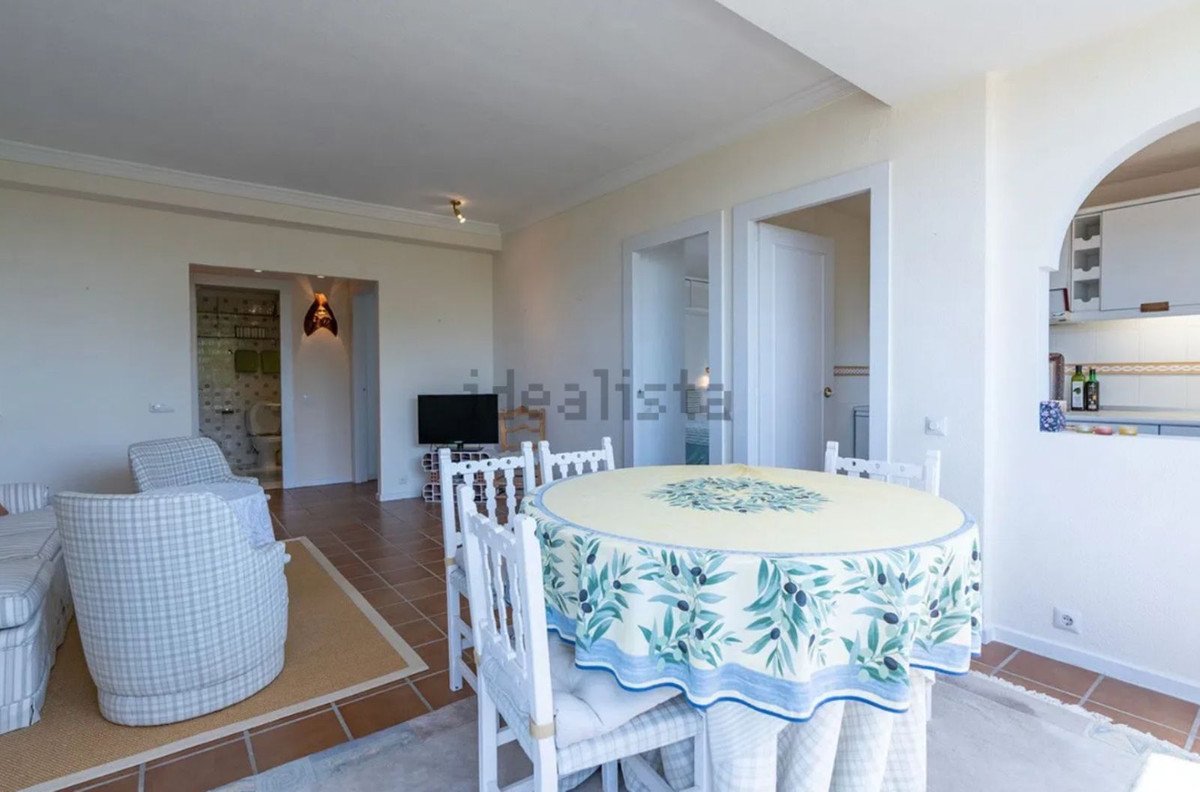 A round dining table with a patterned tablecloth is set in a living area with seating, a TV, and access to a bathroom and kitchen.