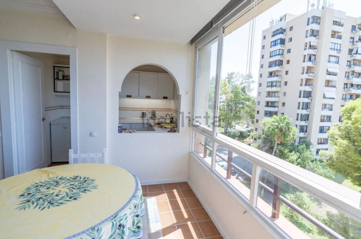 Sunroom with a kitchenette and a table, overlooking a building and greenery from the balcony.